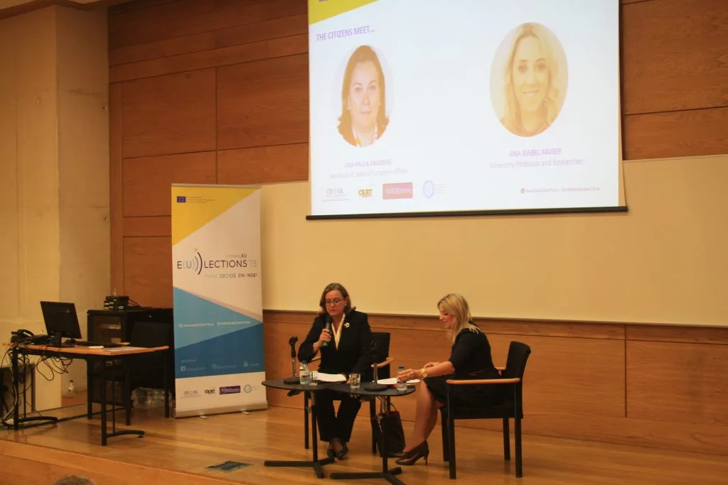 Ana Paula Zacarias and the host seated on the stage during the talk, with its poster projected in the back, containing their pictures and names.