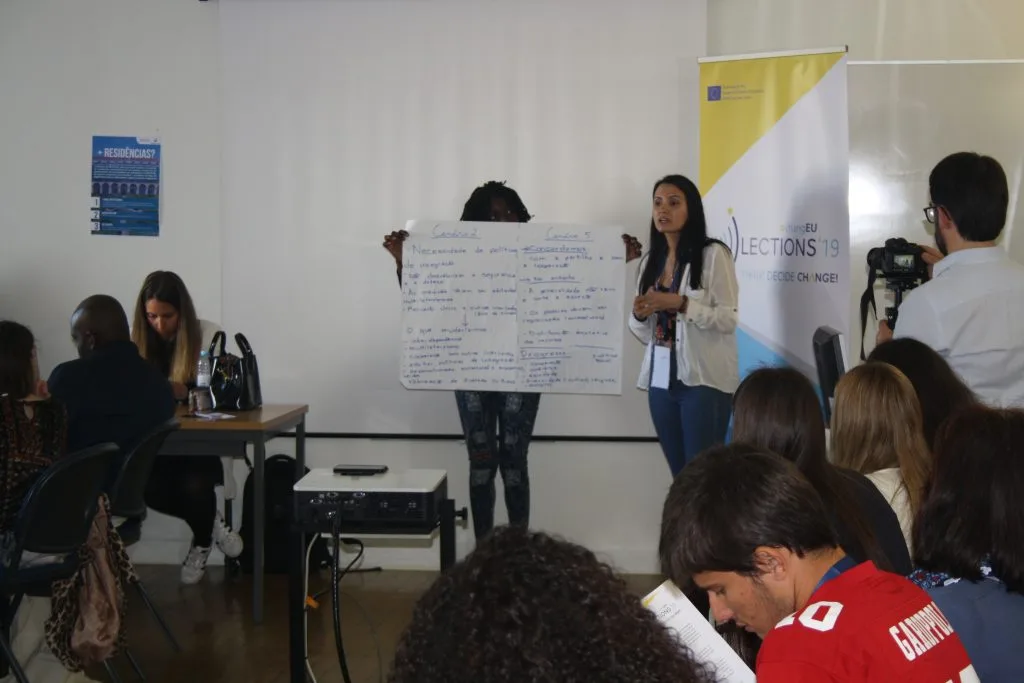 Two students presenting a poster with answers, with one talking while the other is holding it to be seen by the other participants in the classroom.