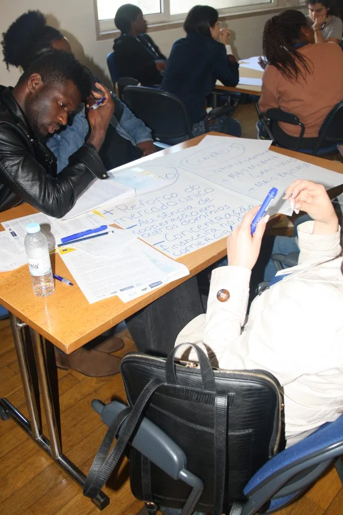 Group of students seated around a table with several papers spread over it, working on their handwritten poster containing the answers.