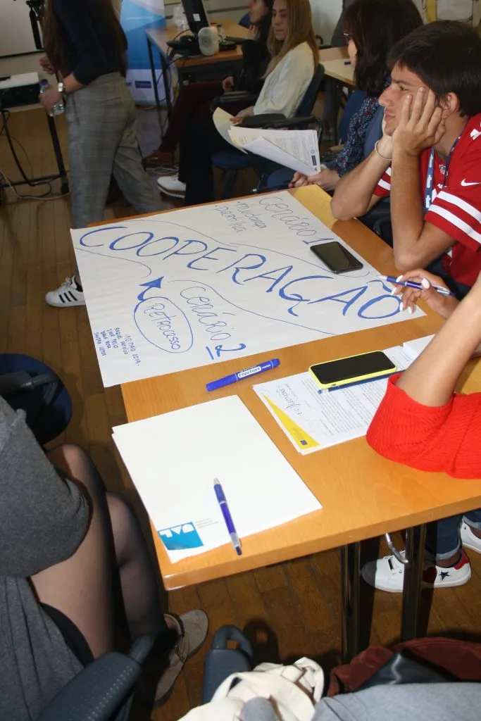 Handwritten poster with the word "cooperation" in portuguese standing out, on a table with three seated participants around it.