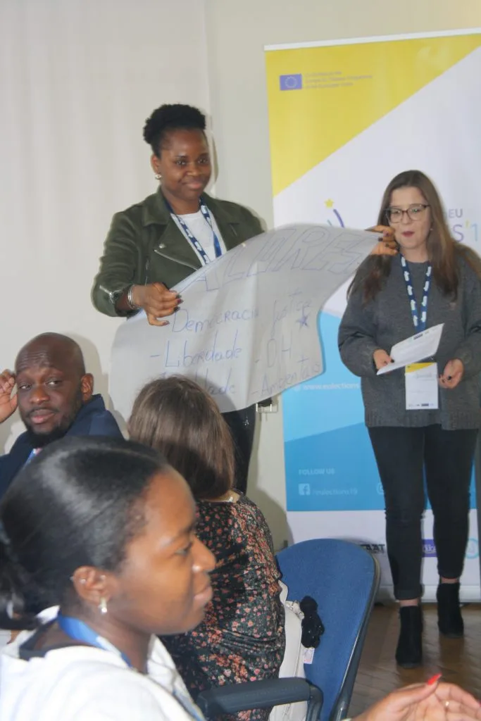 Two participants presenting their poster, with one holding it and the other talking, and a project logo banner behind them.
