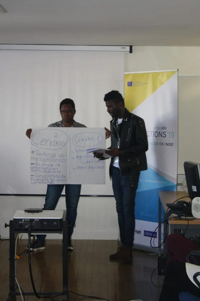Two participants presenting their poster, with one holding it and the other talking, and a project logo banner behind them.