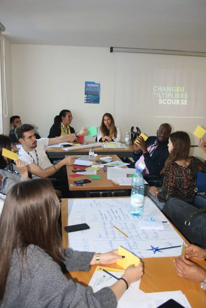 Participants seated around tables, with the colored cards and paper sheets spread over them, while some participants hold multiple cards. A projected slide showing the words "ChangEU Multipliers Webcourse" can be seen in the back.