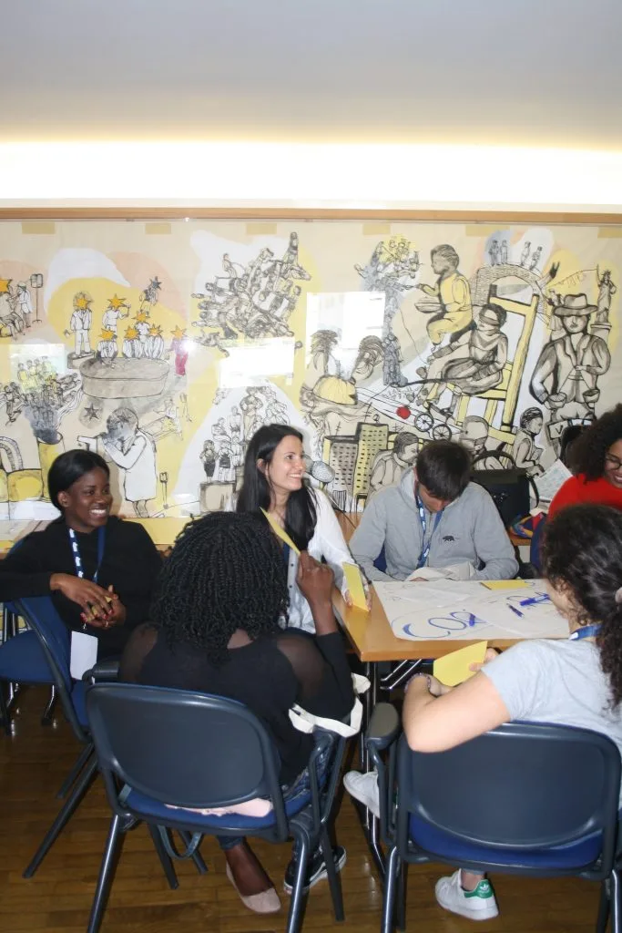 Participants and representatives seated around a table with a handwritten poster over it and some yellow cards, with some people holding them.