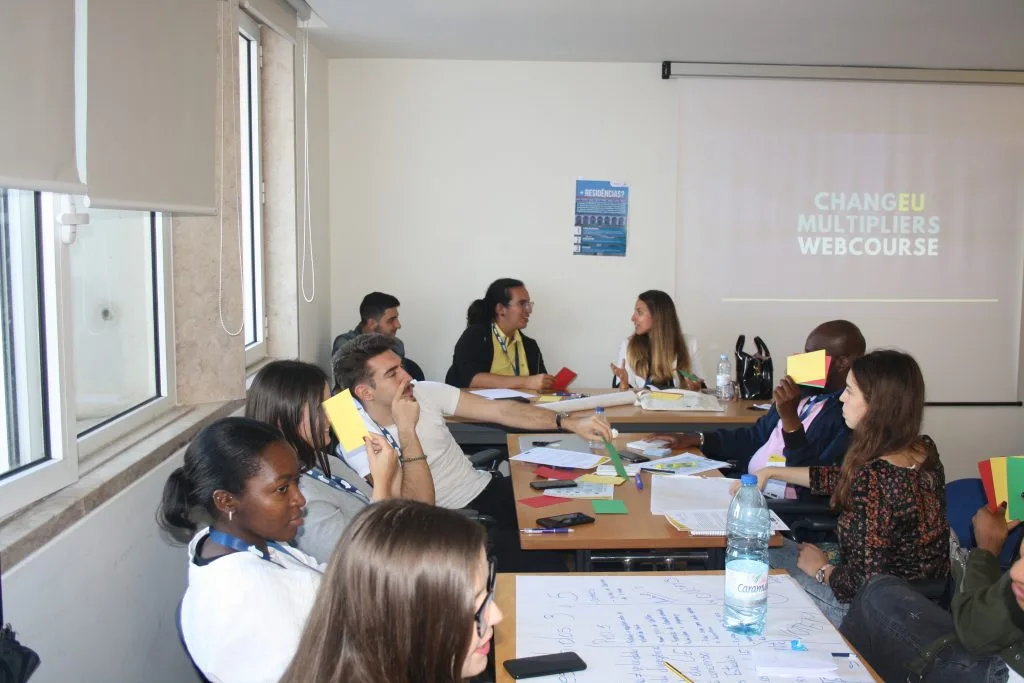 Participants seated around tables, with the colored cards and paper sheets spread over them, while some participants hold multiple cards. A projected slide showing the words "ChangEU Multipliers Webcourse" can be seen in the back.