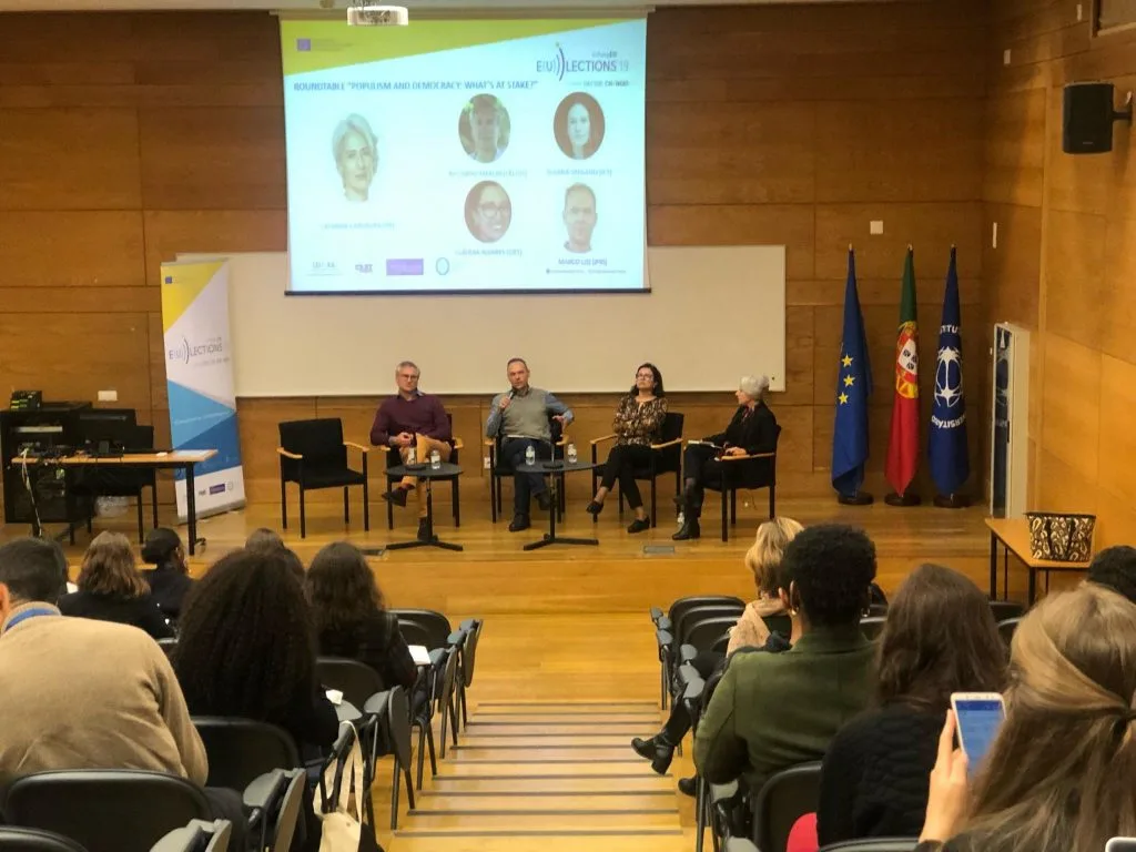 Four roundtable participants seated on stage in front of the auditorium's audience with the event poster projected behind them, perspective from the middle of the audience.