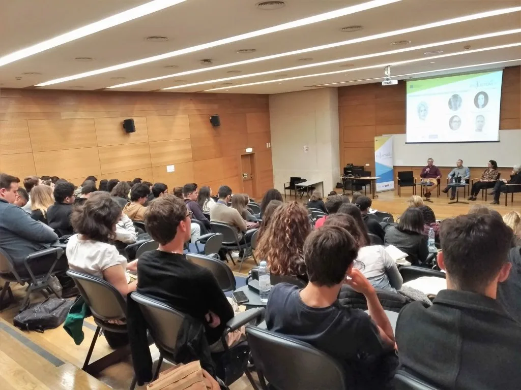 Four roundtable participants seated on stage in front of the auditorium's audience with the event poster projected behind them, perspective from the back of the audience.