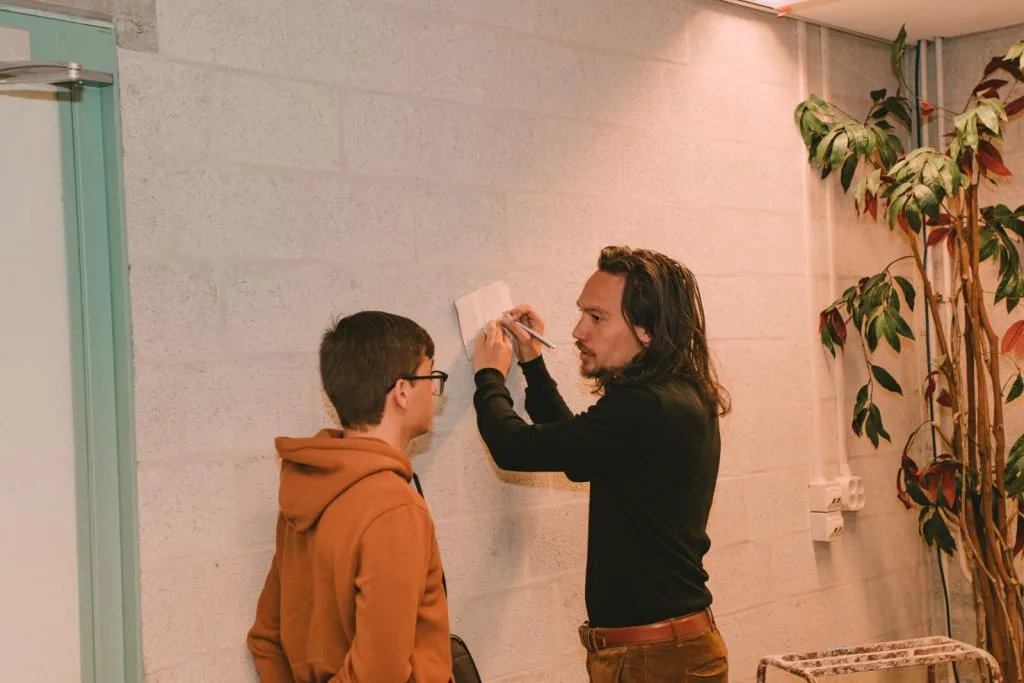 Two participants chatting, one man writing on paper against a wall.