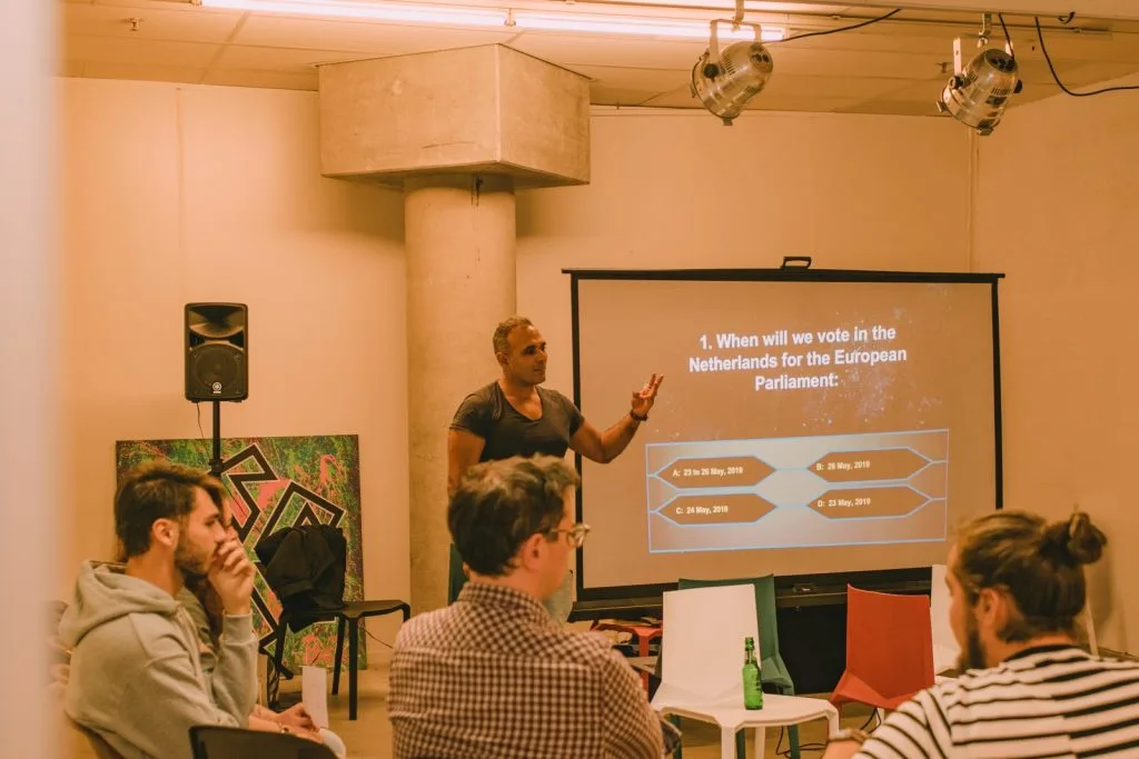 Man presenting slideshow in front of three audience members. Projected image showing "When will we vote in Netherlands for the European Parliament?"