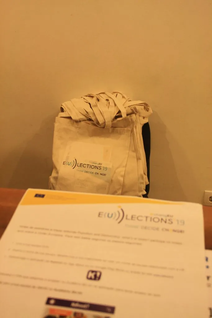 Multiple tote bags with the project logo piled on a chair and a piece of paper on a table in front where the project logo can also be seen.