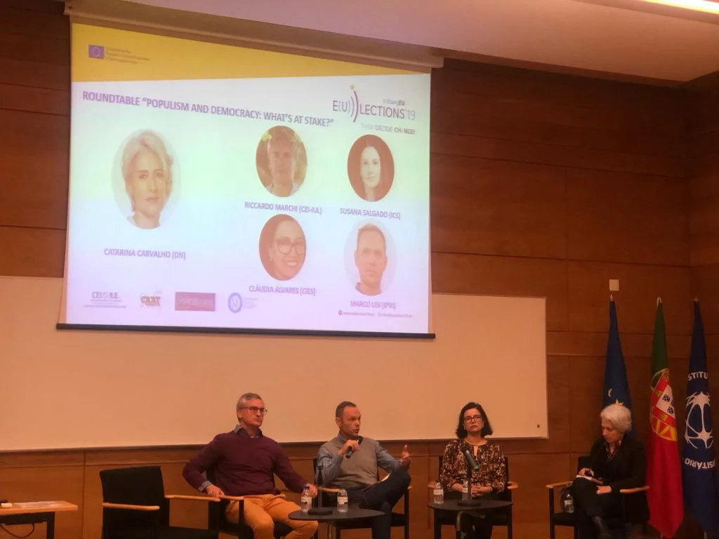 Four roundtable participants seated on stage during the event, with its poster projected behind them.
