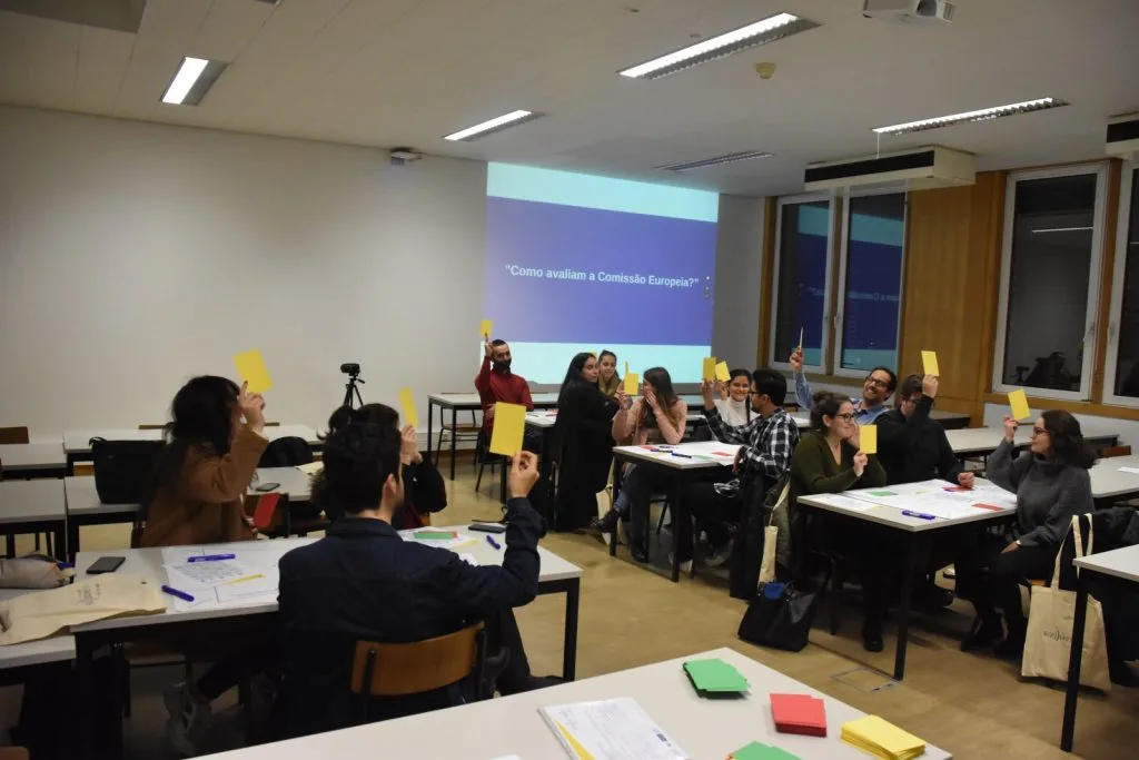 Seated participants holding out yellow cards with the words "How do you evaluate the European Comission?" in portuguese projected on the back.