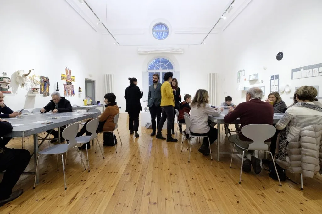 Perspective of the room, with two tables with seated participants on the left and right and more people standing in the center.