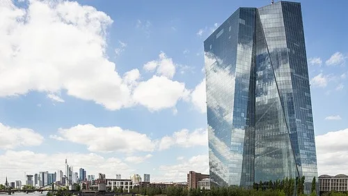 A view of the building of the European Central Bank in Frankfurt Am Main, Germany. A tall building covered in glass windows appears to hover over the city landscape against the sky.