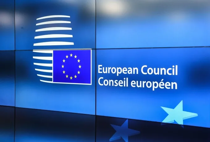 The text "European Council Conseil européen" besides the European Union flag over a blue background, shown on multiple flat screens.