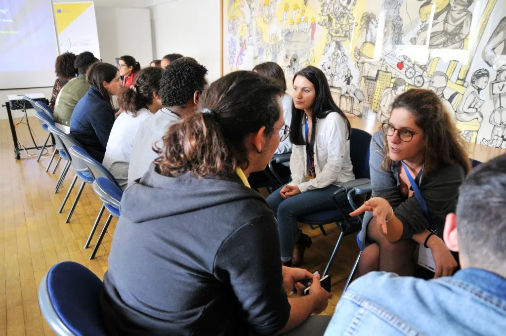 Students seated in two rows, one facing the other, each talking with the person in front of them.