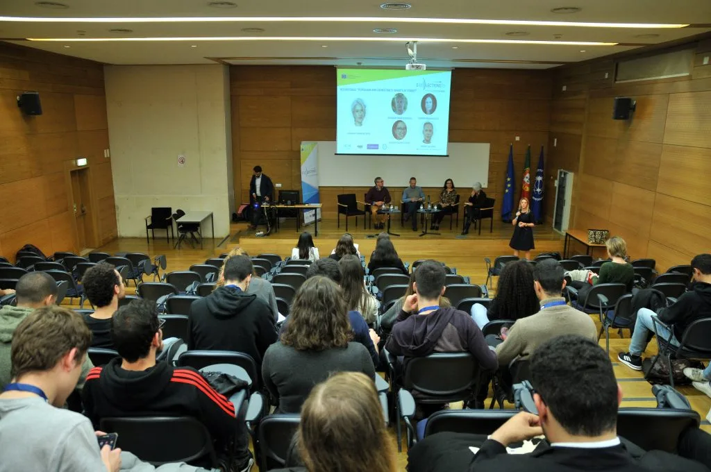 Four roundtable participants seated on stage in front of the auditorium's audience with the event poster projected behind them, with other representative on the left of the stage and the host standing and talking on the right. Perspective from the back of the audience.