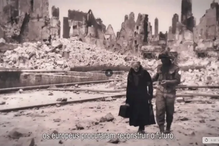Video still with a picture of a destroyed city in the Second World War with an elderly woman and a soldier walking on the right.