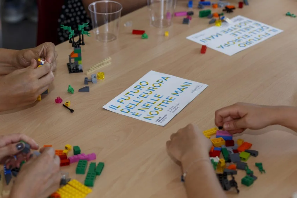 Three participant's hands engaged in making Lego constructions with paper sheets spread over the table containing the phrase "Il Futuro Dell' Europa É Nelle Nostre Mani".