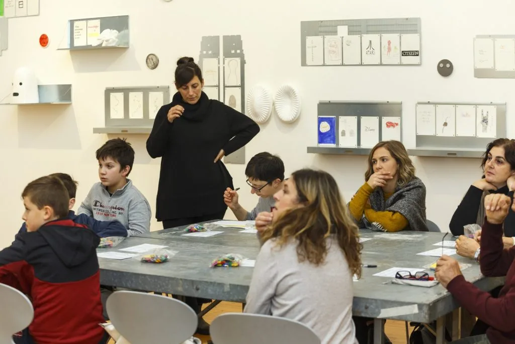 Participating children and adults seated around a table with bags with Lego pieces and paper sheets in front of each one, and a representative standing beside a participant.