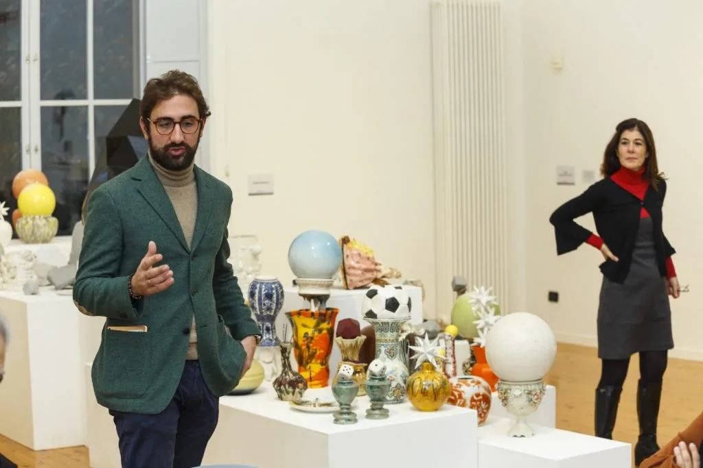 Two representatives speaking, one in front on the left and the other on the right in the back, with several various decorative objects on exposition in the center.