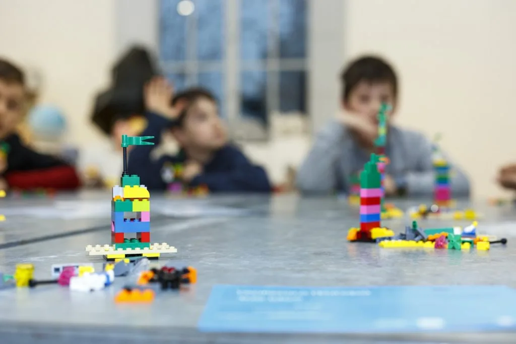 Various Lego constructs across a table with out of focus children in the back.
