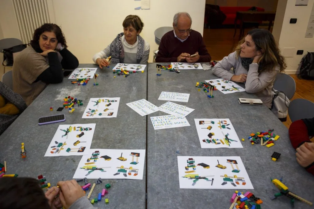 Participants seated around a table constructing with their Lego pieces, with paper sheets showing examples in front of them.