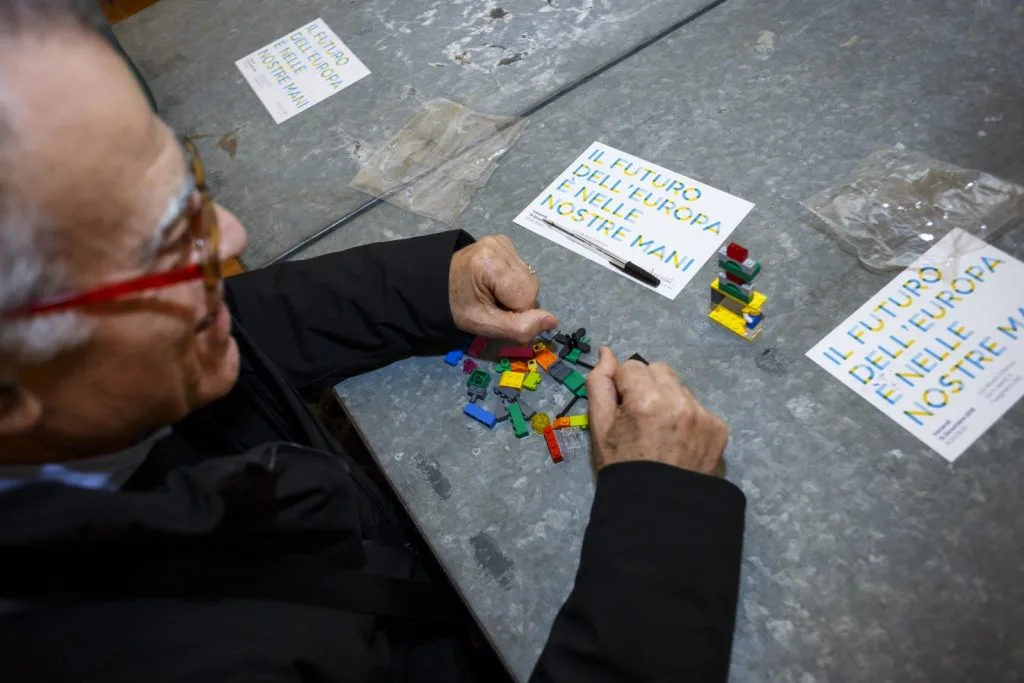 Participant constructing with Lego pieces and three papers with "Il Future Dell' Europa É Nelle Nostre Mani" in front of them.