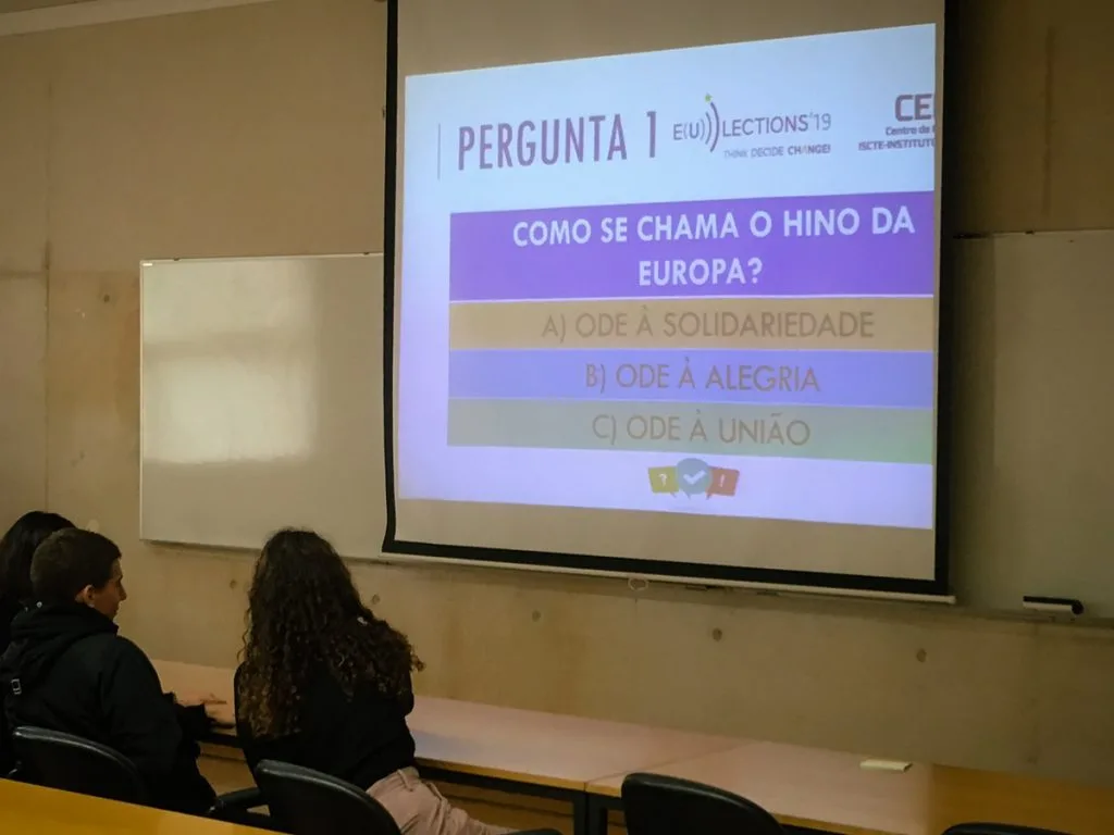 Projected slide containing the question "What is the name of the hymn of Europe?" in portuguese, with two students seated in front.